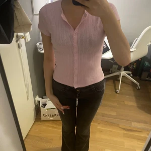 Rosa ribbad tröja från Hollister - Supersöt ljusrosa tröja från Hollister med korta ärmar och knappar hela vägen framtill. Toppen är ribbad med diskret hålmönster och har en liten broderad logga på bröstet. Perfekt för dig som gillar en feminin och fräsch stil. Materialet är tunt och väldigt skönt! Super bra till sommaren eller våren. Helt ny har aldrig använt den men har redan klippt prislappen.