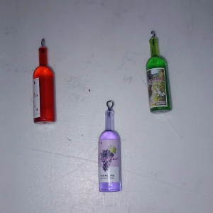 Färgglada vinflaska-halsband - Tre halsband med hängen formade som vinflaskor i rött, grönt och lila. Varje flaska har en etikett med olika motiv och färger. Perfekt accessoar för dig som gillar unika och lekfulla smycken. Hängena är gjorda i plast med metallögla för kedja.