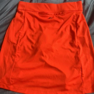 Orange sportkjol från J.Lindeberg XS - Snygg orange sportkjol från J.Lindeberg i stretchigt material. Kjolen är kort och har inbyggda shorts under, perfekt för aktiva dagar. Diskret logga på baksidan och bred midjeresår för extra komfort.