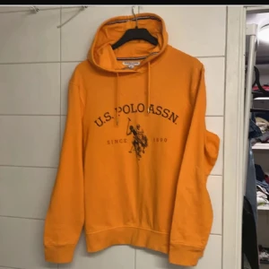 Orange hoodie från U.S. Polo Assn. - Snygg orange hoodie från U.S. Polo Assn. med svart logga och tryck framtill. Tröjan har huva med snörning och ribbade muddar. Perfekt för dig som gillar sportig och avslappnad stil.