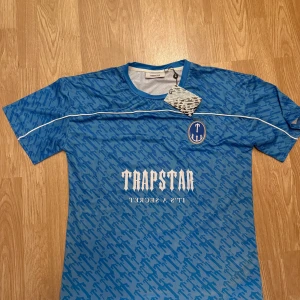 Blå Trapstar fotbollströja - Snygg blå fotbollströja från Trapstar med vit logga och text på bröstet samt siffran 22 på ryggen. Tröjan har ett randigt mönster i blå toner, korta ärmar och vita detaljer längs axlarna. Perfekt för dig som vill sticka ut på planen eller gatan.