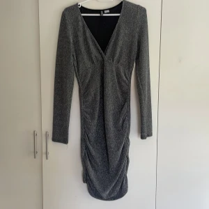Glittrig kortklänning från H&M  - Snygg glittrig kortklänning från H&M Divided i svart och silver. Klänningen har v-ringning, långa ärmar och rynkade detaljer längs sidorna. Perfekt för dig som vill sticka ut med en festlig look!
