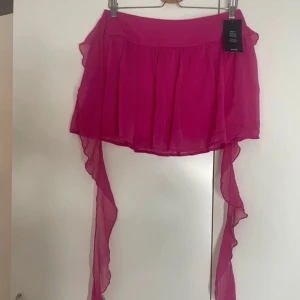 Rosa volangkjol från NA-KD - Supersöt rosa kortkjol från NA-KD med asymmetrisk design och långa volangdetaljer på sidorna. Kjolen är luftig och har en flowy känsla, perfekt för att sticka ut. Färgen är starkt rosa och materialet känns lätt och somrigt.