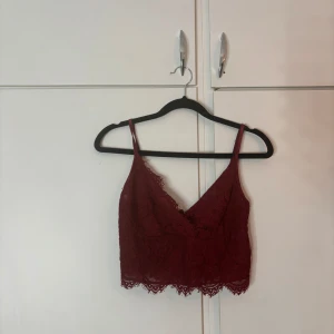 Röd festtopp - Säljer en vinröd croppad linnetopp köpt på New Yorker med blommig spets och smala axelband. Toppen har v-ringning och dragkedja i ryggen för en snygg detalj. Storlek M men den är ganska liten i storlek så skulle säga att det mer är en XS eller S. Använd några fåtal gåner