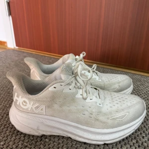 Vita Hoka Clifton 9 skor - Säljer ett par vita Hoka Clifton X sneakers med tjock, dämpad sula och ventilerande mesh-ovandel. Skorna har snörning och en sportig, modern look med stora HOKA-loggor på sidan. Perfekta för dig som gillar bekväma och snygga träningsskor. Lite smutsiga men knappt använda, köpta ett halv år sen. Strl 41, men sitter som 40.