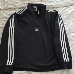 Svart Adidas half zip sweatshirt M - Svart Adidas sweatshirt med klassiska vita ränder längs ärmarna och broderad logga på bröstet. Half zip framtill och hög krage ger en sportig vibe. Tillverkad i mjuk bomullsmix, perfekt för chill eller träning.