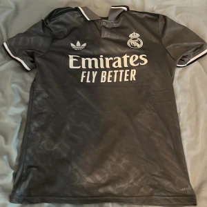 Real Madrid Mbappé Adidas fotbollströja - Snygg mörkgrå Real Madrid fotbollströja med Mbappé och nummer 9 på ryggen. Tröjan har klassisk krage, vita detaljer och broderade loggor från Adidas och Real Madrid. Tillverkad i lätt och ventilerande polyester, perfekt för match eller träning.