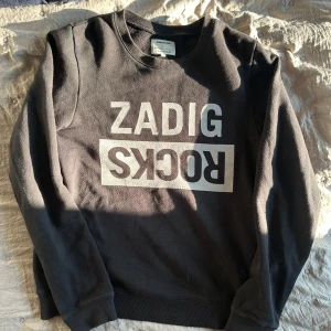Svart sweatshirt från Zadig & Voltaire - Cool svart sweatshirt från Zadig & Voltaire med stor vit logga framtill. Klassisk rund halsringning och långa ärmar. Mjuk bomullskvalitet och ribbade muddar vid ärmslut och nederkant. Perfekt för en avslappnad och trendig look.