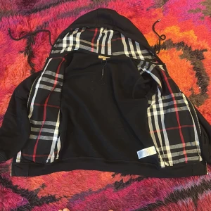 Svart Burberry vindjacka med rutig huva - Svart vindjacka från Burberry med klassiskt rutigt foder i huvan och insidan. Jackan har dragkedja, snörning i huvan och är tillverkad i bomull. Perfekt för dig som vill ha en stilren look med en touch av lyxig streetstyle.