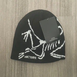 Svart mössa Arc'teryx med vit logga - Svart mössa från Arc'teryx med stor vit dinosaurielogga och broderad text framtill. Snygg, tight passform och tillverkad i mjukt stickat material. Perfekt för kalla dagar när du vill ha både stil och värme.