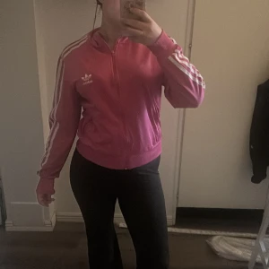 Rosa Adidas track jacket med vita ränder - Säljer en rosa Adidas track jacket med klassiska vita ränder längs ärmarna och stort Adidas-tryck på ryggen. Jackan har dragkedja framtill, ribbade muddar och är i sportig, snygg passform. Perfekt för dig som gillar streetstyle och sportiga vibes.
