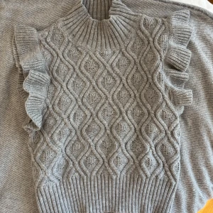 Grå stickad ärmlös tröja från SHEIN - Supersöt grå stickad tröja från SHEIN med hög krage och volangdetaljer på axlarna. Snyggt mönster i garnet och ribbade kanter. Perfekt att styla över en skjorta eller t-shirt för en trendig look.