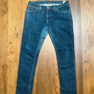 Blå jeans från Nudie Jeans - Snygga blå jeans från Nudie Jeans med klassiska orange sömmar och ikonisk broderad bakficka. Jeansen har normal midja och är tillverkade i mjukt bomullsjeans. Perfekta för dig som gillar en smal passform och stilren look. Storlek 32/32. En defekt vid insida lår men inget som märks vid användning! Längd 102 cm, bredd 41 cm. Fraktar samma dag! 