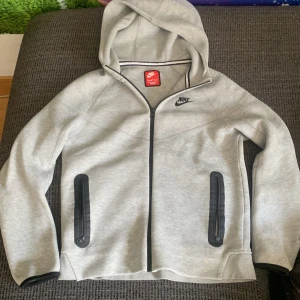 Grå hoodiejacka från Nike med dragkedja - Snygg ljusgrå Nike tech från Nike med hel dragkedja framtill och svarta detaljer vid fickorna. Jackan har huva, långa ärmar och är tillverkad i mjukt material som passar perfekt till chill eller träning. Nike-logga broderad på bröstet.