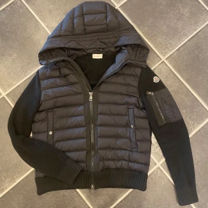 Svart Moncler tricot cardigan - Snygg svart cardigan från Moncler med stickade ärmar och vadderad kropp. Jackan har huva, dragkedja framtill, ribbade muddar och en Moncler-logga på ärmen. Perfekt för dig som vill ha en stilren och sportig look.