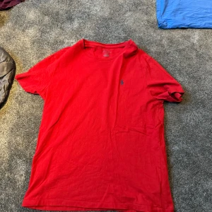 Röd t-shirt från Polo Ralph Lauren - Röd t-shirt från Polo Ralph Lauren med klassisk passform och liten blå broderad logga på bröstet. Tillverkad i mjuk bomull och har rund halsringning samt korta ärmar. Perfekt för en clean och stilren look.