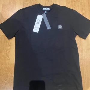 Svart t-shirt från Stone Island - Svart t-shirt från Stone Island med klassisk logopatch på bröstet. T-shirten har rund halsringning och korta ärmar. Tillverkad i mjuk bomull för en skön känsla och clean look. Perfekt för dig som gillar stilrena plagg med streetkänsla.