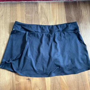 Svart sportkjol från Decathlon - Kort sportkjol från Decathlon i stretchigt syntetmaterial. Kjolen har en enkel design med bred midjeresår och innerbyxa, perfekt för träning eller tennis. 