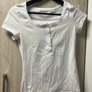 Vit kortärmad topp med knappar - En basic vit topp i bomull med rund halsringning och korta ärmar. Framsidan har en rad med sex vita knappar som ger en enkel men snygg detalj. Perfekt att matcha med jeans eller kjol för en clean look. Aldrig använd 