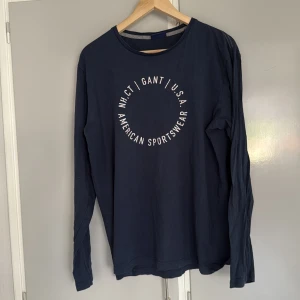 Marinblå långärmad tröja från Gant Medium - Snygg marinblå långärmad tröja från Gant med vit tryckt text i cirkel på bröstet. Tröjan har en klassisk rund halsringning och är gjord i mjuk bomull, blekare än ny.