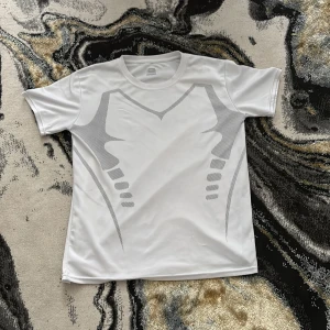 Vit sportig t-shirt XL - Vit t-shirt i sportig stil med grått grafiskt mönster framtill. Tillverkad i ett lätt och svalt syntetmaterial som passar perfekt för träning. Klassisk rund halsringning och korta ärmar för en clean look.