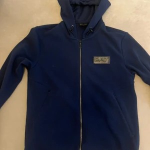 Blå EA7 hoodiejacka med dragkedja - Snygg marinblå hoodiejacka från EA7 Emporio Armani med dragkedja framtill och två fickor. Ändå skavanken är att märktet har missfärgats i tvätt. Nypris 2200kr. S men lite större i storleken.