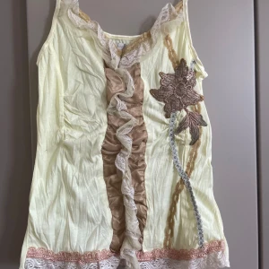 Ljusgul linnetopp med spets från Saint Tropez - Supersöt ljusgul linnetopp från Saint Tropez med bred spetskant nertill och runt halsen. Framsidan har en beige volang med spetsdetaljer och en broderad blomma i brunt och beige. Toppen är ärmlös och har en romantisk, bohemisk vibe.