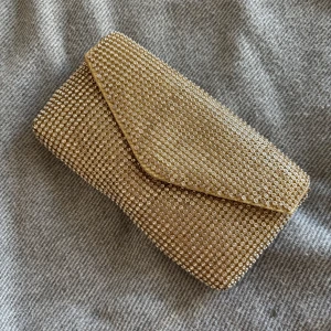Glittrig guldig kuvertväska med kedja - Säljer en glittrig kuvertväska i guld med massor av små pärlor och en avtagbar guldfärgad kedja. Väskan har magnetlås och insida i ljus satin. Perfekt när du vill sticka ut och addera extra bling till din outfit.