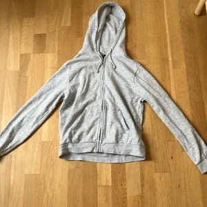 Ljusgrå hoodie från SOC med dragkedja - Ljusgrå hoodie från SOC i storlek S. Tröjan har huva med snörning, hel dragkedja framtill och två fickor. Tillverkad i mjuk bomullsblandning, perfekt för chill eller träning. Klassisk och enkel design som funkar till det mesta. Tröjan har dock en väldig liten fläck (vilket man kan se på sista bilden).