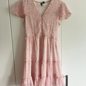Ljusrosa blommig klänning Vero Moda XS - Supersöt ljusrosa klänning från Vero Moda med små blommor i rosa, v-ringning och knappar framtill. Volangdetaljer och korta ärmar ger en romantisk vibe. Perfekt för varma dagar och somriga tillfällen.