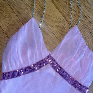 Ljusrosa långklänning med paljettdetalj - Superfin ljusrosa långklänning med glittriga smala axelband och v-ringning. Klänningen har en markerad midja med rosa paljetter och ett flowigt, transparent yttertyg. Perfekt för dig som vill sticka ut med en feminin och glamorös look.