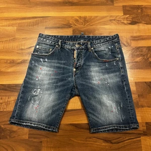 Dsquared2 jeansshorts med färgstänk - Snygga jeansshorts från Dsquared2 i mörkblå denim med slitningar och coola färgstänk i rött och vitt. Klassisk femficksmodell med gylf och knapp, samt Dsquared2-logga på bakfickan. Perfekta för dig som gillar streetstyle och vill sticka ut.
