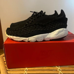 Nike Air Footscape Woven NM svart/vit - Nike Air Footscape Woven NM sneakers i svart med unik flätad ovandel och vit chunky sula. Skorna har snörning och en modern, sportig look som sticker ut. Materialmix av textil och mocka ger en cool känsla och komfort. Perfekta för dig som gillar statement sneakers.