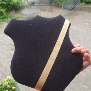 Svart smyckeshållare med guldfärgat band - En stilren smyckeshållare i svart tyg med ett diagonalt guldfärgat band. Perfekt för att visa upp halsband eller kedjor. Sköldformad design som enkelt kan hängas upp på väggen tack vare metallfäste på baksidan.