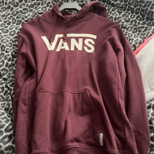 Vinröd hoodie från Vans - Snygg vinröd hoodie från Vans med stor vit logga på bröstet. Tröjan har huva med snörning och en klassisk känguruficka framtill. 