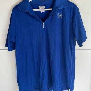 Blå pikétröja med strassblomma - Blå pikétröja med kort ärm och klassisk krage. Halv dragkedja framtill och en snygg blomma i strass på bröstet som ger lite extra bling. Perfekt för dig som gillar sportig men ändå söt stil.