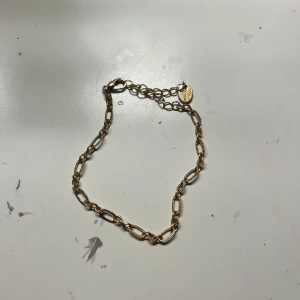 Guldigt kedjearmband med detalj - Snyggt armband i guldton med ovala länkar och en liten bricka vid låset. Kedjan har en cool, klassisk design som passar perfekt till din vardagsstil. Justerbar längd med karbinhake för enkel användning.
