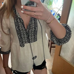 Vit blus med svart brodyr och knyt - Supersöt vit blus med svart broderat mönster längs v-ringningen och ärmsluten. Blusen har trekvartsärmar, knytband med tofsar och pärldetaljer. Luftig och flowy passform, perfekt för vår och sommar. Materialet känns som bomull och ger en bohemisk vibe.