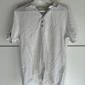 Vit t-shirt med knappar och krage - Vit t-shirt med korta ärmar, liten krage och knappslå med fyra knappar framtill. Tillverkad i mjuk bomull, perfekt för en clean och enkel stil. Passar dig som gillar klassiska plagg med en twist.