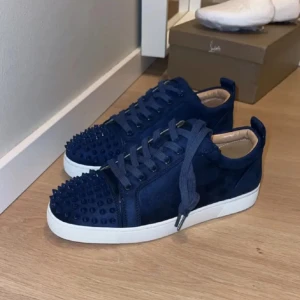 Mörkblå sneakers från Christian Louboutin - Snygga mörkblå sneakers i mocka från Christian Louboutin med ikoniska röda sulor och coola nitar på tån. Skorna har snörning och vit platt sula, perfekt för dig som vill sticka ut med en lyxig och edgy stil.