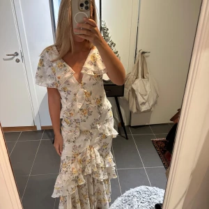 Vit blommig långklänning med volanger - Storlek 38 men passar mig som har XS/S i vanliga fall! Superfin långklänning i vit bas med blommigt mönster i gult och lila. Klänningen har v-ringning, volangdetaljer längs hela klänningen och korta ärmar. Perfekt för sommarens alla tillfällen, riktigt romantisk och flowy känsla.