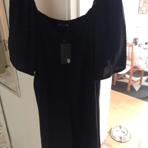 Svart offshoulder klänning 157 - Svart offshoulder klänning från 157 med smockad överdel och vida korta ärmar. Klänningen har en enkel och stilren design, perfekt för dig som gillar en klassisk look med modern twist. Materialet känns lätt och bekvämt mot huden.