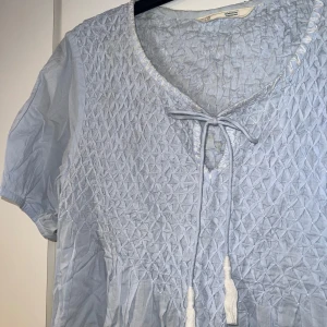 Ljusblå blus från odd Molly - Supersöt ljusblå blus från Odd molly med smockad överdel, knytband med tofsar vid halsen och korta ärmar. Tillverkad i tunn bomull, perfekt för varma dagar. Snygg struktur och avslappnad passform. Passar M-2XL. Aldrig använd. Nypris ca 1300kr 