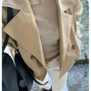 Kort trenchcoat  - Säljer en så snygg kort trenchcoat i beige med stora slag och dubbelknäppning. Jackan har breda ärmar och coola knappar framtill. Perfekt för dig som vill ha en trendig och avslappnad look. Passar lika bra till jeans som till kjol. Använd 1-2ggr i våras