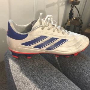 Adidas Copa Pure fotbollsskor vit/blå - Säljer ett par Adidas Copa Pure fotbollsskor i vitt med blå detaljer och röda inslag. Skorna har klassiska tre ränder på sidan, snörning och dobbar för gräsplan. Perfekta för dig som vill sticka ut på planen med stilren design och bra grepp.