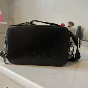 Svart axelväska från Valentino - Snygg svart axelväska från Valentino i skinn. Väskan har två stora fack med silvriga dragkedjor och en bred, justerbar axelrem med Valentino-tryck. Perfekt storlek för det viktigaste. Köpt för 1200kr. Däremot har lite silvriga färg gått bort på ena dragkedjan💕 Skriv för mer info!