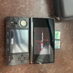 Nitendo 3DS - Bilderna visar en Nintendo 3DS-spelkonsol med tillhörande fodral och spelkassetter, bland annat Super Mario Bros. Perfekt för dig som gillar portabel gaming och klassiska Nintendo-spel. Konsolen ser ut att vara i bra skick och är redo för nya äventyr