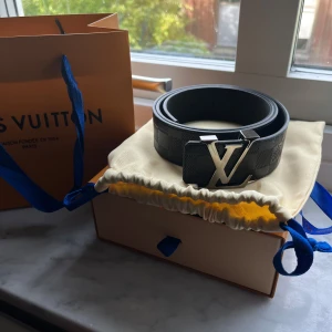 Louis Vuitton Bälte (nyskick) - Säljer ett svart bälte från Louis Vuitton med ikoniskt LV-spänne i svart metall. Bältet har diskret rutmönster och levereras i original ask, dustbag och påse med blå detaljer. Perfekt accessoar för att lyfta vilken outfit som helst. Använd fåtal gånger. Märks inte att den är använd. 110 cm. Köpt från Stockx. Vid snabbaffär kan priset diskuteras. Hör av er vid intresse! 