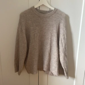 Beige stickad tröja från 157 - Mysig beige stickad tröja från 157 med rund halsringning och långa ärmar. Perfekt för lager-på-lager och chill dagar. Tröjan har en avslappnad passform och ribbade muddar vid ärmslut och nederkant. Enkel att matcha med jeans eller kjol.