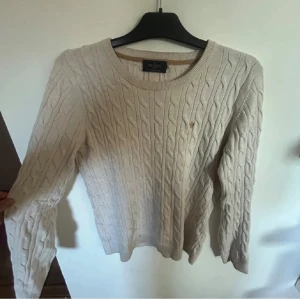 Beige kabelstickad tröja Morris - Kabelstickad beige tröja från Morris med rund halsringning och diskret broderad logga på bröstet. Tröjan har lång ärm och klassisk passform, perfekt för dig som gillar en stilren och tidlös look. Materialet är mjukt och känns skönt mot huden.